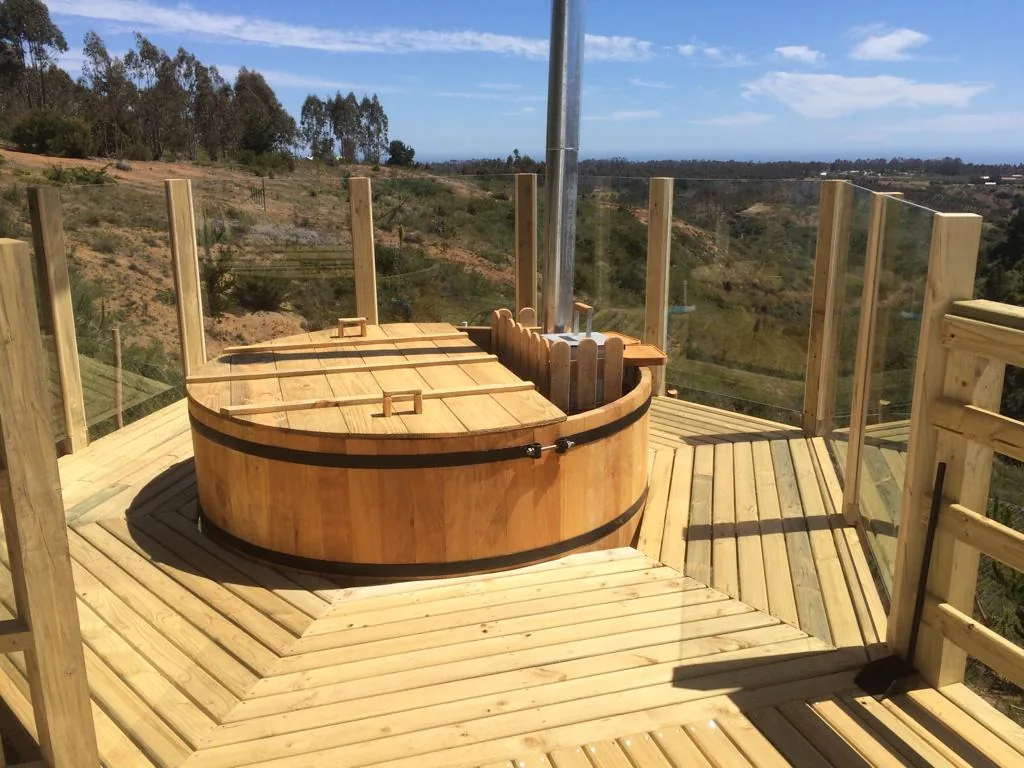 Tina de madera circular con chimenea sobre un deck con baranda de vidrio y vista panorámica al valle y el mar